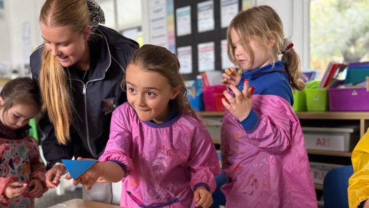 Photo of 3 female kindergarten students wearing bright pink paint smocks with a female teacherher