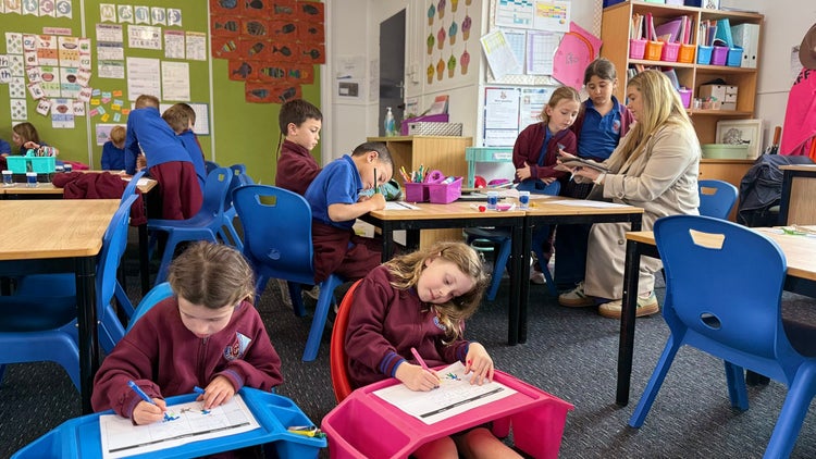 Photo of 2 female Year 1 students sitting on the floor with a female teacher and other students working