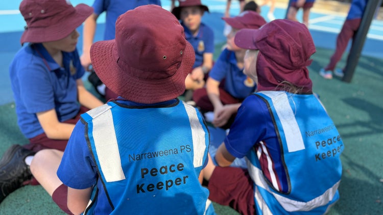 Photo of male Year 5 students sitting in playground with 2 students wearing blue high vis vest labelled Peace Keeper