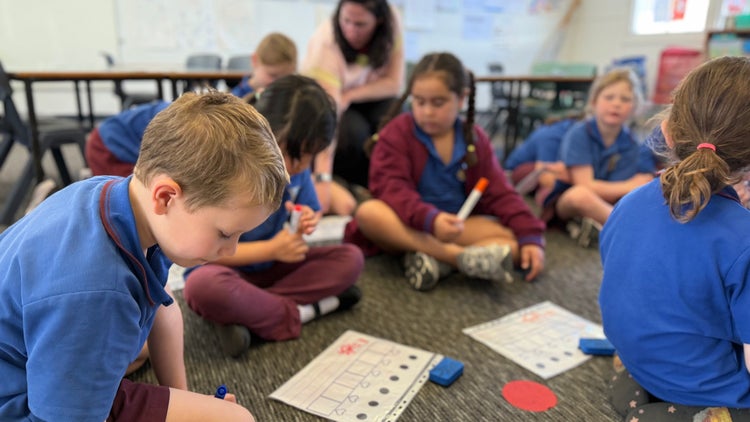 Photo of male and female kindergarten students sitting on the floor using mini whiteboards with teacher in background