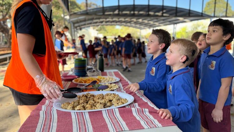 Photo of a table with fruit and muffins and male Year 1 students lining up to be served afternoon tea by female educator wearing a high vis vest